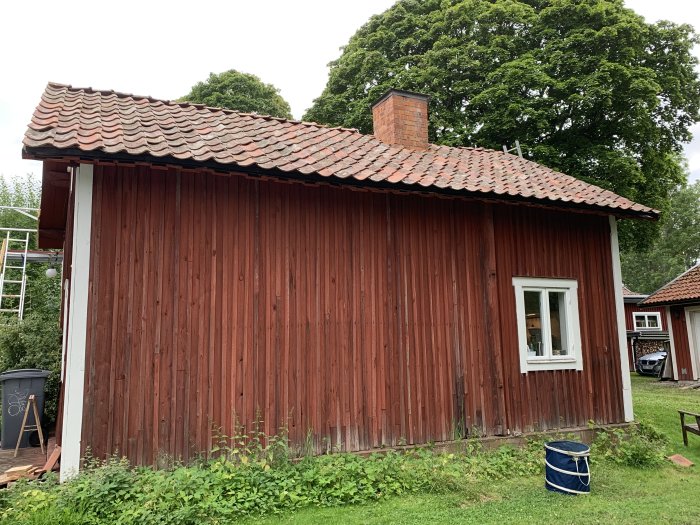 Ett rött soldattorp med vitkantade fönster och tegeltak, omgivet av grönska och en stege lutad mot taket.