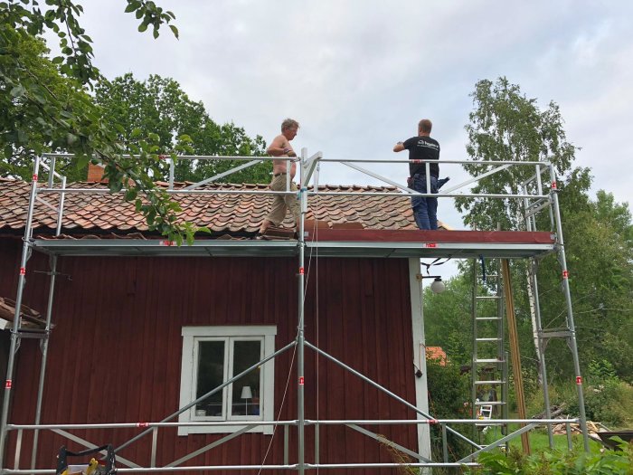 Två personer på en byggnadsställning inspekterar taket på ett traditionellt rött trähus.