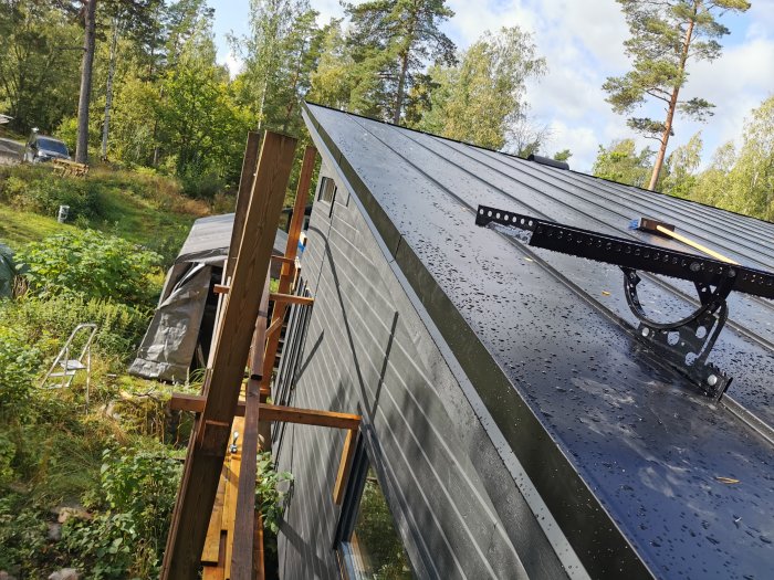 Pågående bandtäckningsarbete på hus med skivtak, ställningar och natur i bakgrunden.