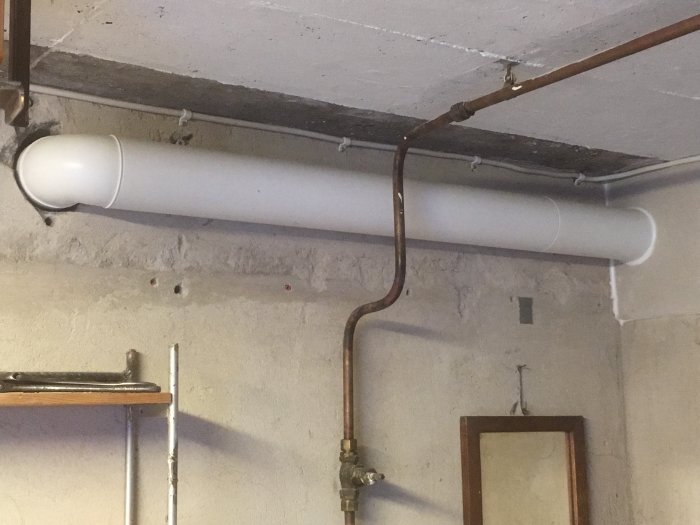 Nyinstallerad vit ventilationskanal i plast längs en källarväggs betongyta med synliga rör och kablar.
