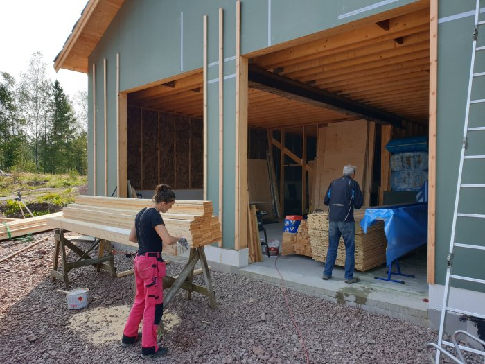 Två personer arbetar med byggnadsmaterial vid en nybyggd struktur omgiven av grus och grönska.