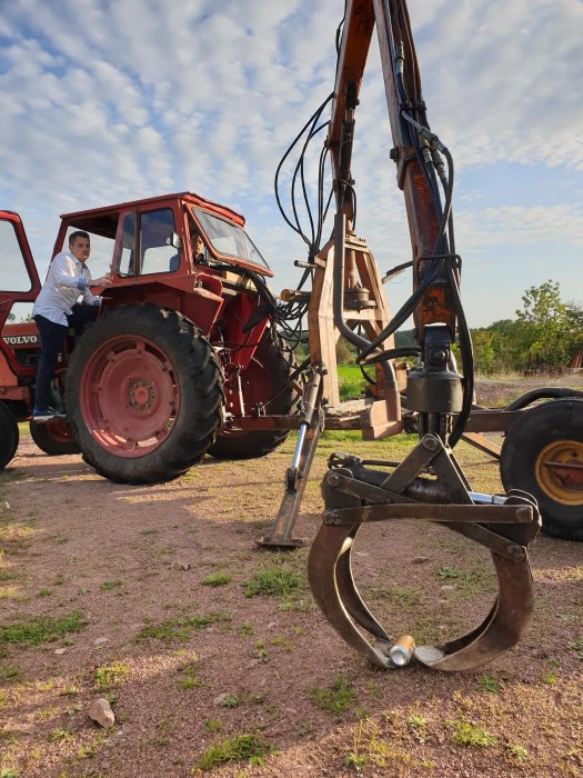 Person står vid en röd traktor med tillkopplad skogskran, klar för tävling i flytta ölburk.