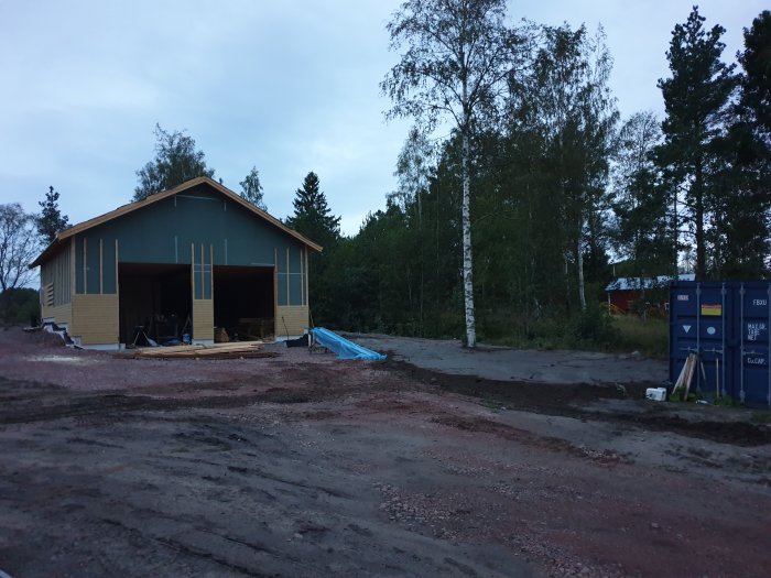 Ett nybyggt hus med öppna portar och byggmaterial framför, på en nylagd grund med skymningen som bakgrund.