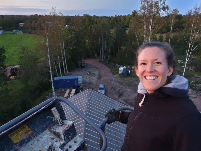 Kvinna i skylift ler mot kameran med byggplats och gröna fält i bakgrunden vid skymning.