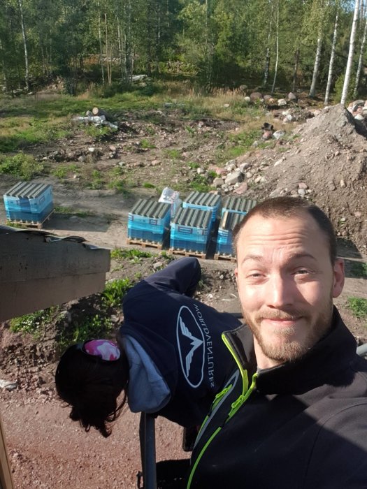 Man som tar en selfie med en byggarbetsplats i bakgrunden, inklusive högar av material och en del grönska.