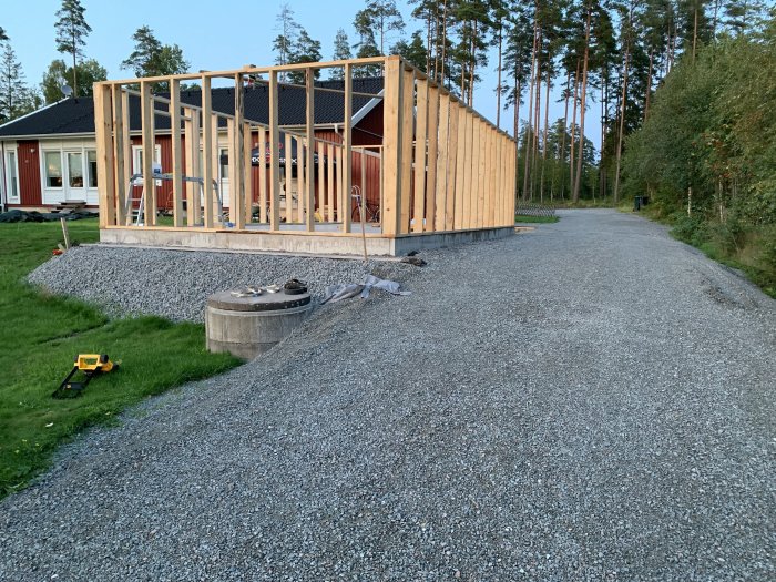 Byggplats med en delvis konstruerad trästomme för ett hus, grusgrund, spräckt sten och byggmaterial, i ett skogsområde vid skymning.