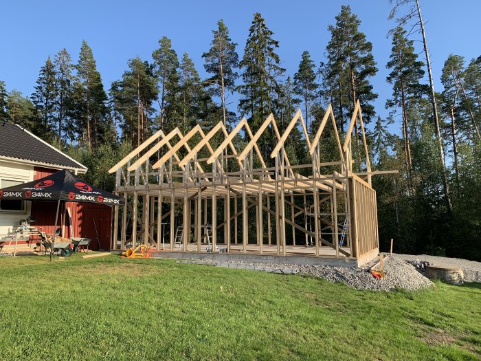 Nybyggd trästomme för hus på betongplatta, gräsmatta i förgrunden och skog i bakgrunden.