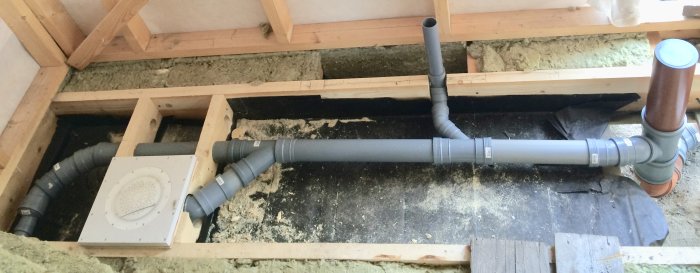Öppet golv med plastmatta och synlig rörinstallation, kortlingar och en avloppsanordning i ett pågående byggprojekt.