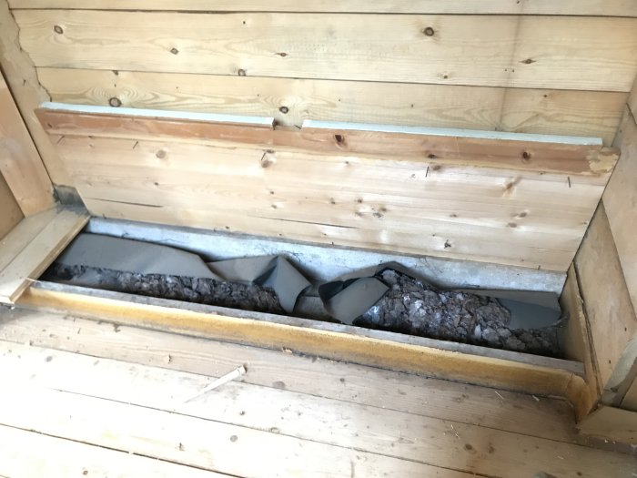 Golvsektion i äldre hus med brunt material, som kan vara isolering, på svart papp under trägolv.