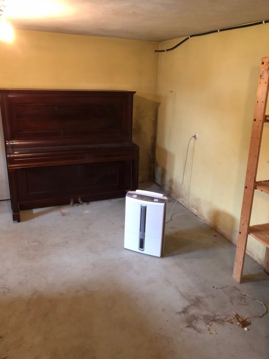 Oinredd källargillestuga med betongväggar i gulaktig ton och ett lackat betonggolv i grön färg. Möbler inkluderar ett brunt piano och trähylla.