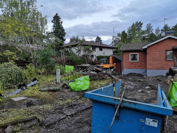 Trädgård under renovering med grävmaskin, sprängt berg, en blå container och en husfasad i bakgrunden.