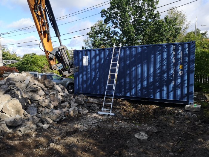 Blå container placeras på byggarbetsplats med berg och grushög, grävmaskinsarm synlig, stege lutad mot containern.