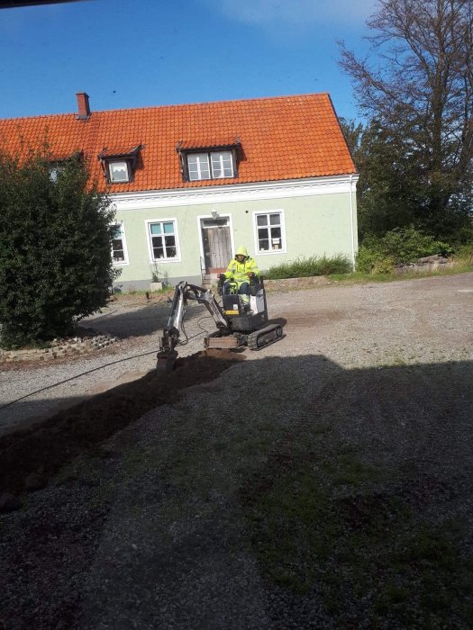 En person i säkerhetskläder gräver med en grävmaskin framför ett hus med orange tak.