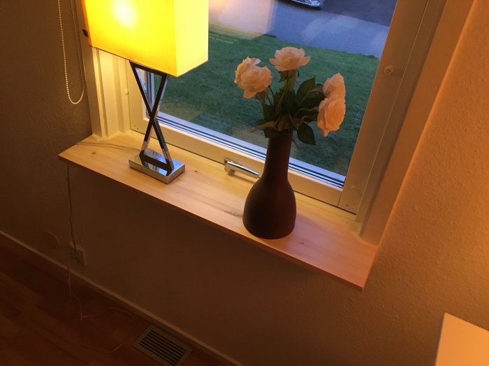 Nyinstallerad obehandlad furufönsterbänk med vas och blommor under en tänd lampa.