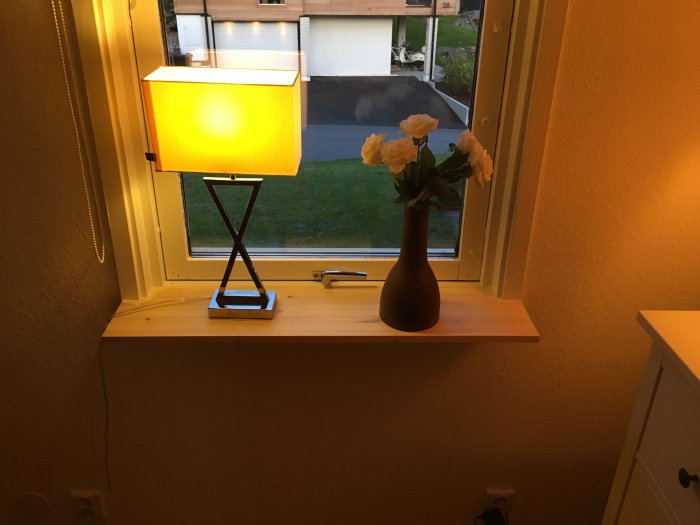 Obehandlad fönsterbänk i furu med lampa och vas med vita blommor i ett hem.