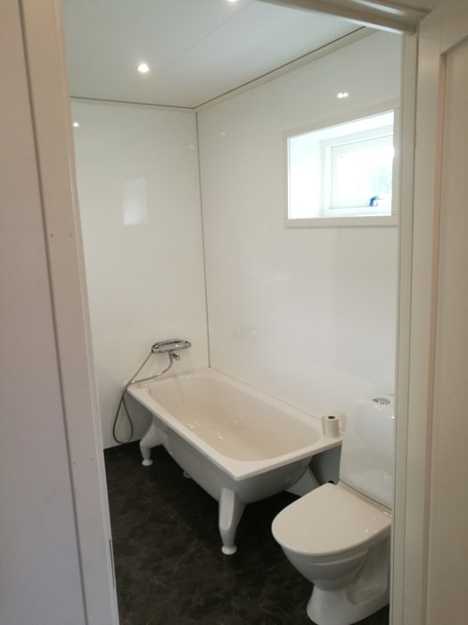 Ett nytt badrum med vit badkar och toalett, mörkt golv och vita väggar med infällt fönster.