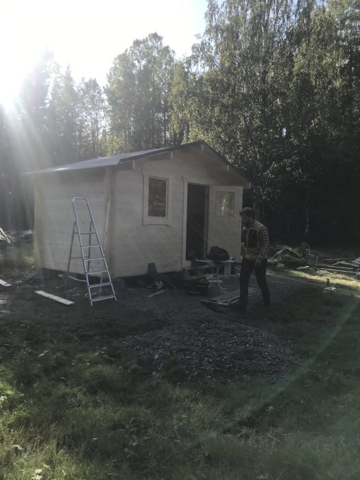 Person framför nyligen påbörjat byggprojekt av liten stuga med underlagspapp på taket och byggmaterial runtomkring.