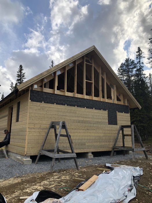 Halvfärdigt trähus med takstolar och delvis påsatt träpanel under byggnation mot skogsbakgrund.