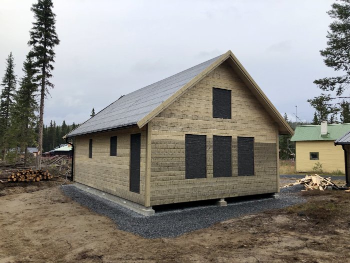 Nybyggd trästuga med spånbelagd fasad och klart underlagstak, omgiven av skogsmark och singelgrund.