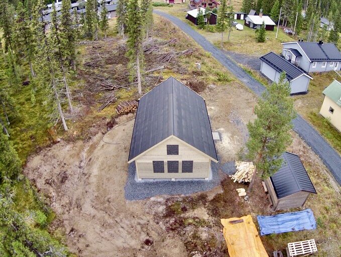 Flygbild av en nybyggd stuga med mörk takbeklädnad i en skogsglänta, omgiven av grusväg och mindre byggnad.