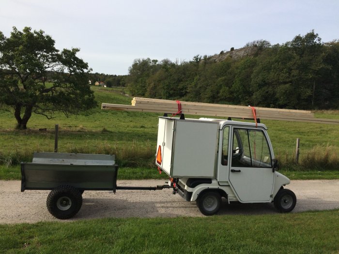 Vit liten lastbil med släpkärra, för långa träreglar fastsatta på taket, står på en lantlig väg.