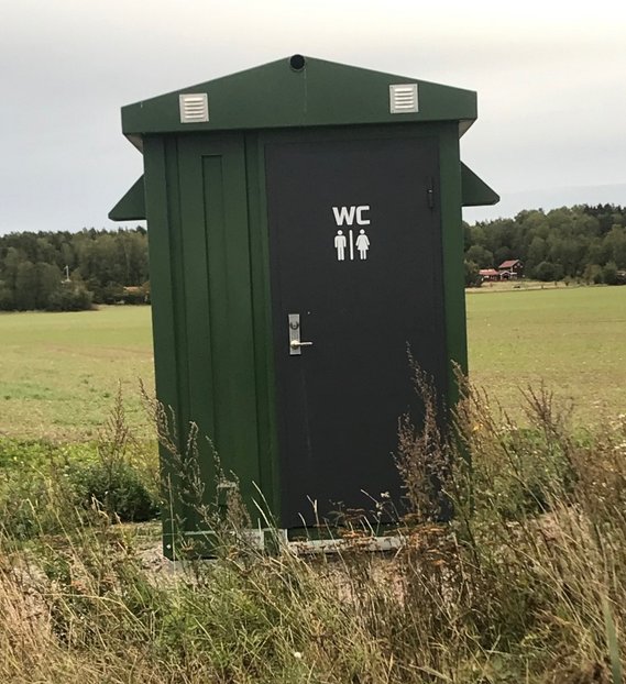 Grönt "kopplingshus" på en äng med WC-symboler sprejade på dörren, vilket ger sken av en offentlig toalett.
