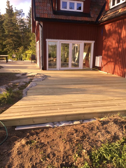 Nybyggd trädäck altan intill rött hus med öppna terrassdörrar och trädgårdsmöbler i bakgrunden.