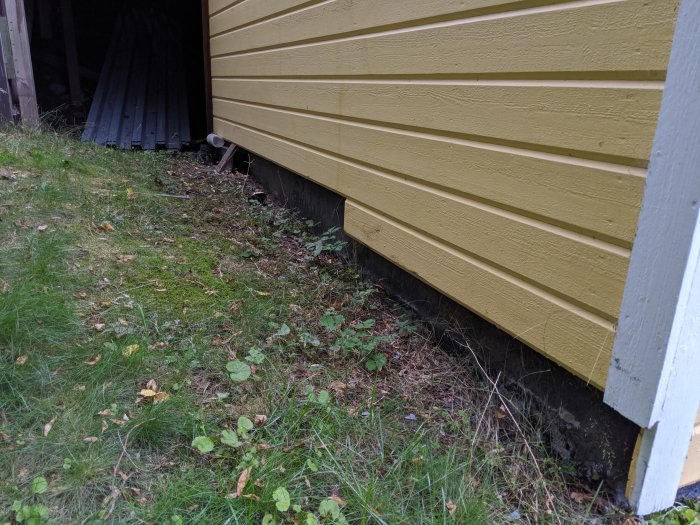 En gul träfasad ovanför en synlig betonggrund på en sluttande tomt, ingen dränering synlig, vegetation nära grunden.