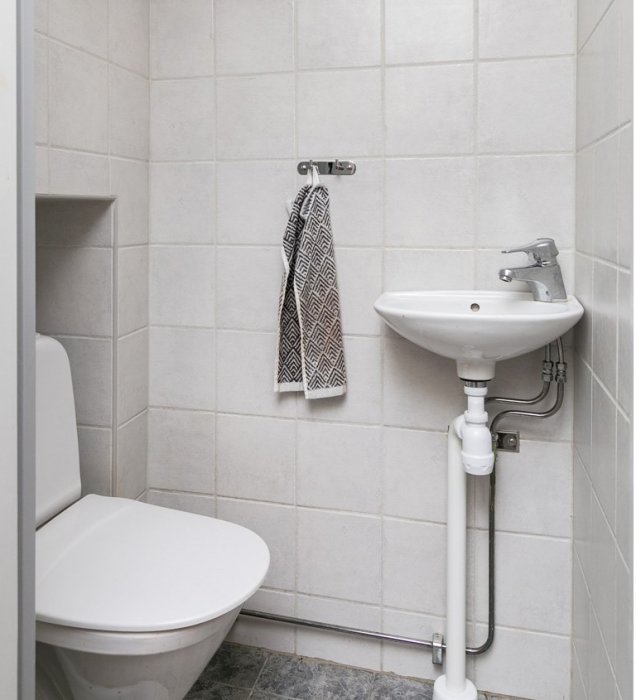 Badrum med toalett och handfat, ingen synlig cistern, kakelväggar och handduk på krok.