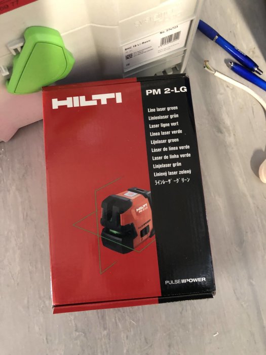 HILTI PM 2-LG linjelaser i förpackning på grått underlag med grönt mäthjälpmedel och blå penna i bakgrunden.