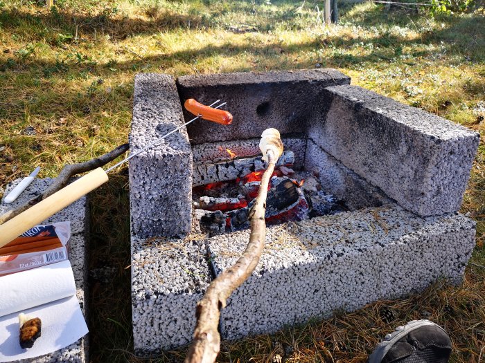 Korv grillas på spett över öppen eld i en enkel, utomhus grillplats av betongblock.