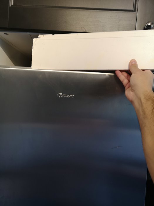 En hånd holder en hvid træplade over et køleskab med et mærke, ved et hulrum under et skab, hvor pladen skal bruges som fyld.