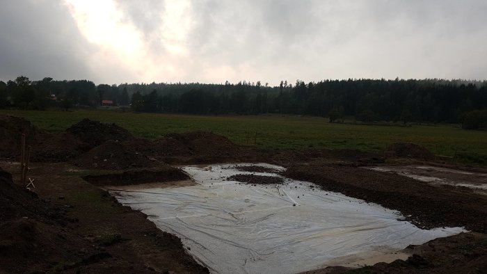 Nygrävd grund med isolering och armering inför gjutning, omgiven av jordhögar mot skogsbakgrund.