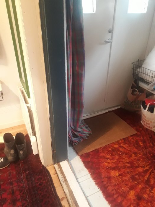 Tröskel vid öppen dörr med röd matta, skor och en färgglad filt i hemmiljö.