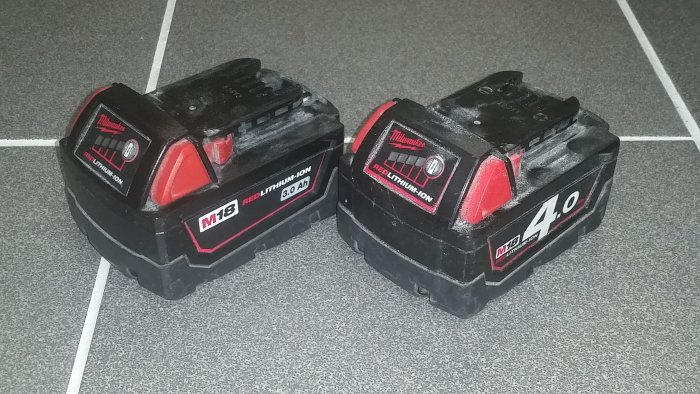 Två svarta Milwaukee M18 RedLithium 3.0Ah och 4.0Ah batteripaket ligger på ett grått klinkergolv.