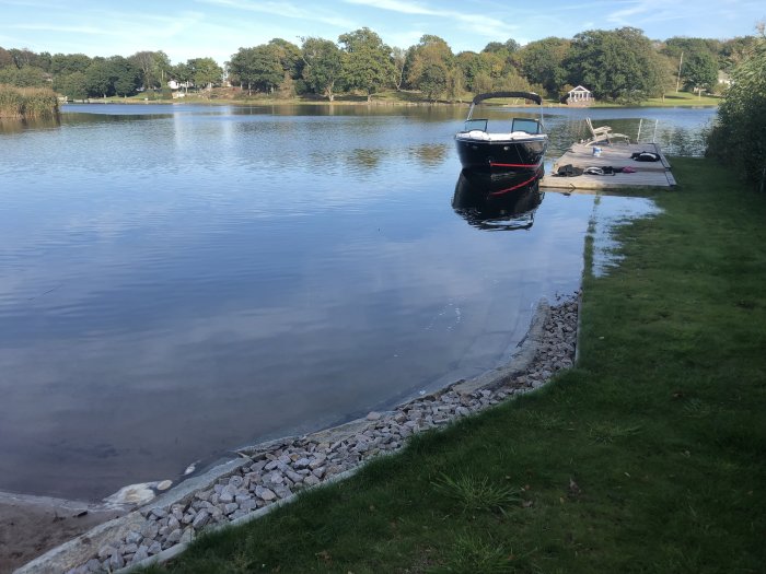 Motorbåt förtöjd vid en liten brygga, med stigande vattennivå nära gräsmatta och stenläggning.