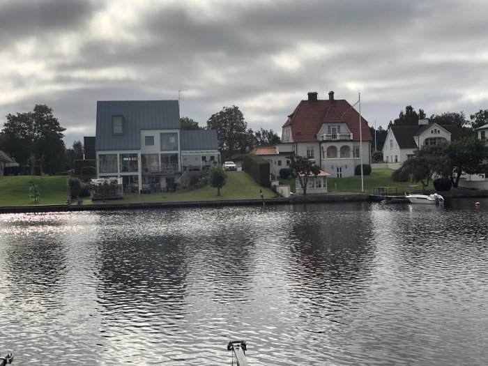 Vy över en lugn sjö mot hus med varierande arkitektur, gröna trädgårdar och en liten båt vid kajen.