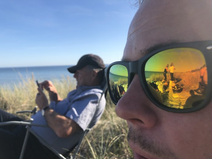 Reflektion av en stranddag med familjen i en mans solglasögon, en annan man i bakgrunden.