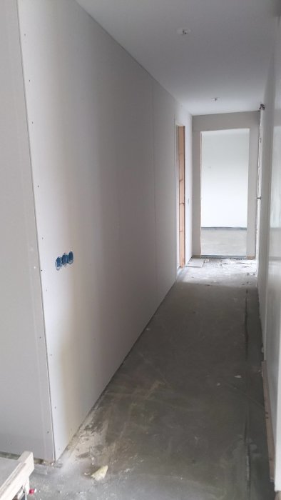 Nybyggd korridor med vita innerväggar, eluttag utanpå vägg och ett betonggolv redo för ytterligare renovering.