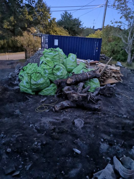 Säckar med rivningsavfall och buntar med virke framför en container.