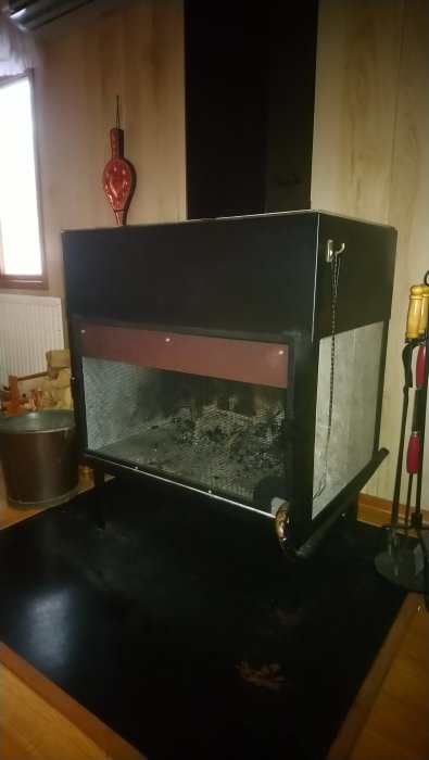 Kamin med öppen spisgrind, vedhink och eldstadsverktyg i ett mörkt rum.