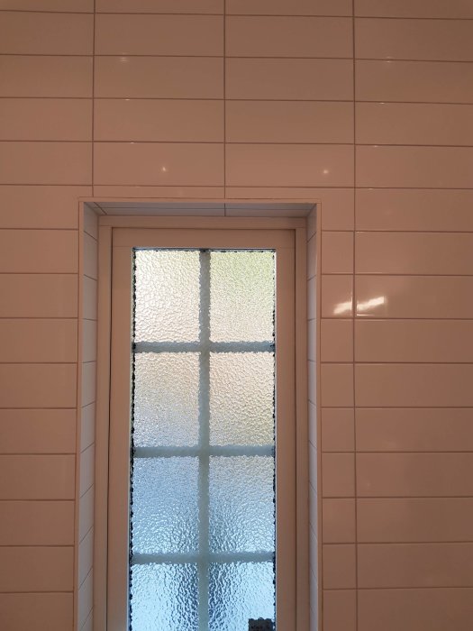 Kaklat badrum med beige kakel symmetriskt runt ett frostat glasfönster, med en önskan om helt symmetrisk placering.