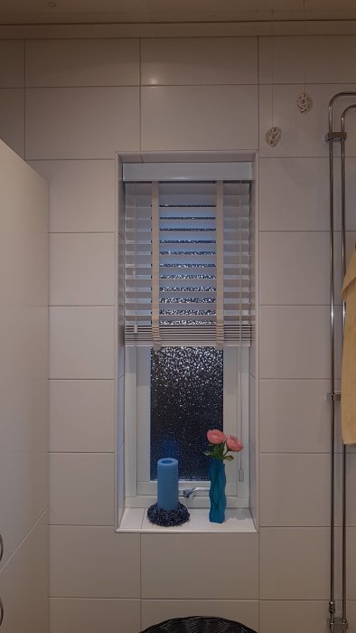 Kakelvägg i badrum med symmetrisk klinkerplacering runt ett fönster med persienner, dekorerat med blå ljus och rosor.