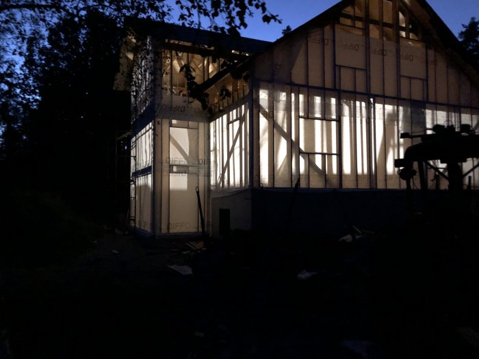 Hus under konstruktion vid skymning med väderskyddande duk och synlig trästomme, innan slutlig fasad är på plats.