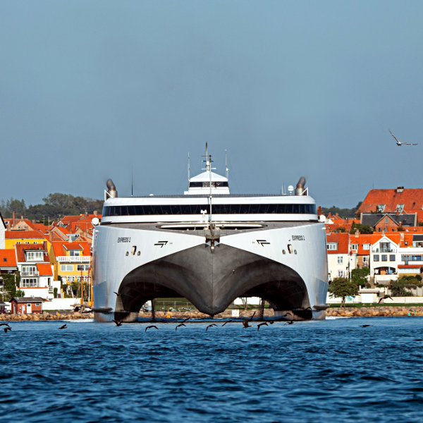 Katamaranfärja framför stad kör i hög fart med bogvågor, sedd framifrån.