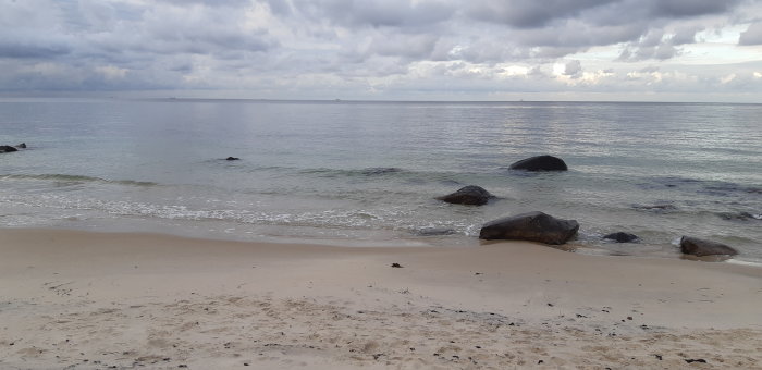 Sandstrand med stenar i vattnet mot bakgrund av en molntäckt himmel.