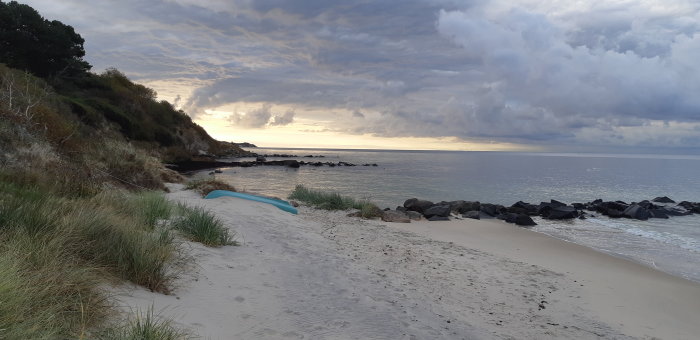 Strand med sanddyner och gräs, omgiven av klippor och träd vid solnedgång.