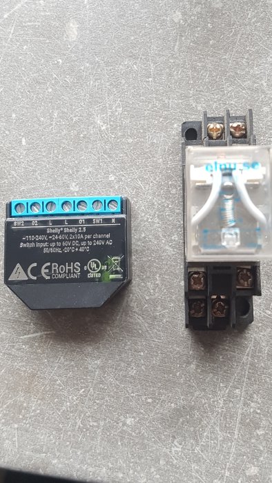 Elektriska installationskomponenter med en svart Shelly 2.5 switch och en genomskinlig timerrelä, ligger på ett grått underlag.