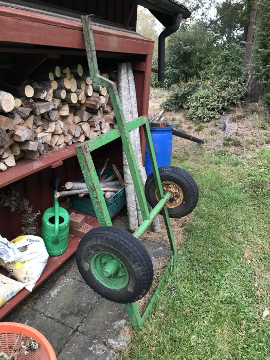 Ett tungt, litet grönt släpvagnsställ med stora hjul står vid en vedbod.