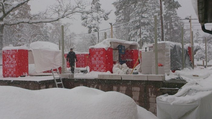 Byggplats under snöfall med lättbetongblock, bjälklag och presenningstak, samt arbetare och utrustning.